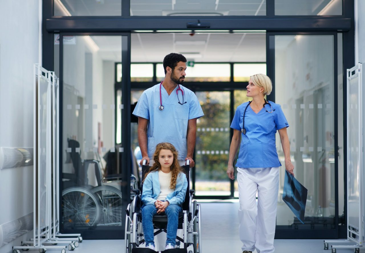 Pregnant doctor with her colleague pushing litle girl at wheelchair at pediatric department.